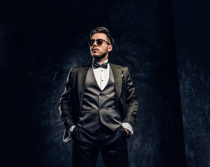 A stylish young man in sunglasses dressed in an elegant suit posing with a hand in a pocket against a dark textured wall