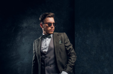 A stylish young man in sunglasses dressed in an elegant suit posing with a hand in a pocket against a dark textured wall