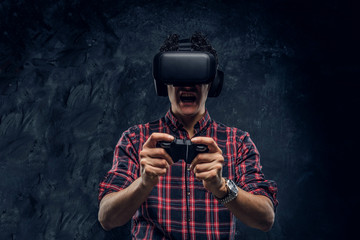 Young emotional man using VR headset and playing a video game with joystick in a studio against the background of the dark wall