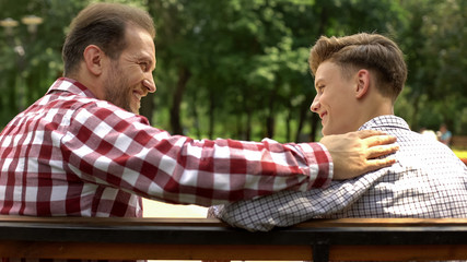 Teenage son talking with father on bench in park, dad supporting his child