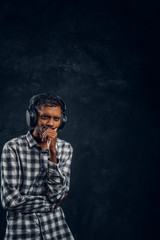 Portrait of a handsome Indian man wearing a checkered shirt listens to music in wireless headphones with thoughtful look standing in studio against the background of the dark wall.