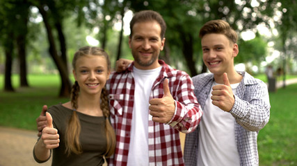 Joyful teenagers and their father showing thumbs up into camera at park, family