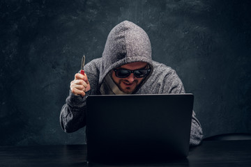 Hacker in hoodie and sunglasses holds a knife lock-up sitting behind a laptop in a dark room