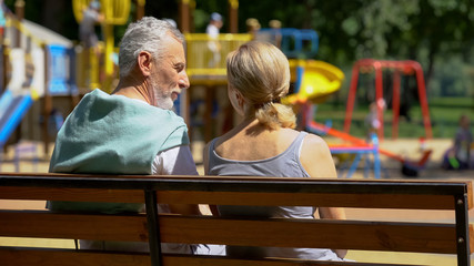 Elderly man and woman communicating on bench in park, watching grandchildren - obrazy, fototapety, plakaty