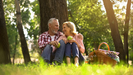 Joyful healthy old couple relaxing on grass, holding apples and hugging, picnic