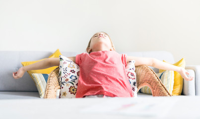 Tired little preschool girl spreading wide arms lying and looking to ceiling in comfortable chair near desk