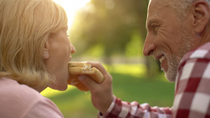 Elderly man feeding his wife fast food burger on picnic, romantic date in park