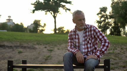 Melancholic elderly male sitting on bench in park and thinking about problems