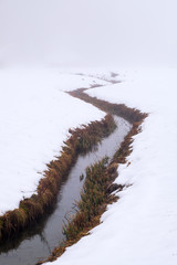 Creek river with bushes on bank winds through snowy field
