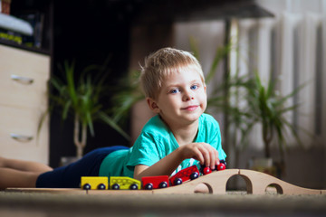 happy baby, blonde with blue eyes, a little boy of five years in the mint (green / blue) t-shirt in shorts barefoot near the couch in the apartment igret fun with wooden railway