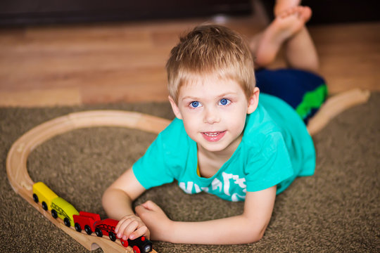 happy baby, blonde with blue eyes, a little boy of five years in the mint (green / blue) t-shirt in shorts barefoot near the couch in the apartment igret fun with wooden railway