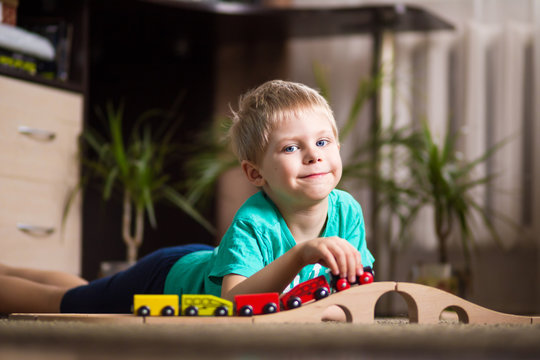 happy baby, blonde with blue eyes, a little boy of five years in the mint (green / blue) t-shirt in shorts barefoot near the couch in the apartment igret fun with wooden railway