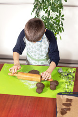 Chłopiec ubrany w fartuch kuchenny wałkuje brązowe ciasto drewnianym wałkiem na silikonowej zielonej stolnicy. Dziecko w kuchni. - obrazy, fototapety, plakaty