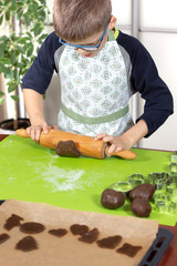 Chłopiec ubrany w fartuch kuchenny wałkuje brązowe ciasto drewnianym wałkiem na silikonowej zielonej stolnicy. Dziecko w kuchni.