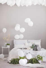White balloons under the ceiling of grey scandinavian bedroom with double bed and green plants in pots, real photo with copy space on the grey wall
