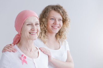 Smiling senior woman suffering from tumor wearing pink headscarf and ribbon, her daughter standing...