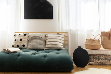 Straw handbags next to stylish black vase and fancy sofa with pillows made of futon, black poster on the wall between windows