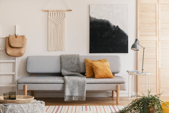 Abstract black and white painting and handmade macrame on white wall of natural living room interior with grey fashionable couch with yellow pillow and warm blanket