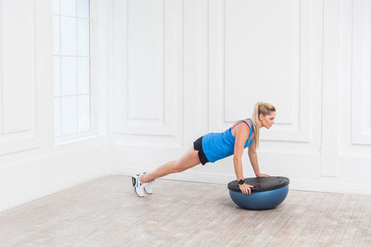 Focused sporty beautiful young athletic blonde woman in black shorts and blue top working in gym doing plank for abdominal muscles on bosu balance trainer, holding balance on fitness ball, indoor