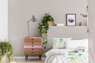 Suede covered pink and olive green nightstand with grey lamp in stylish bedroom with floral sheets and green plants in pots