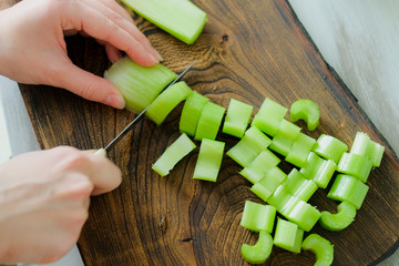 Female hands cut celery stalk with knife on wooden cutting board. Healthy vegan diet