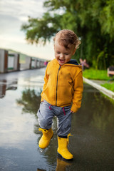 Child in a yellow waterproof coat in the rain jumping in puddles. A boy stands with his head bowed...