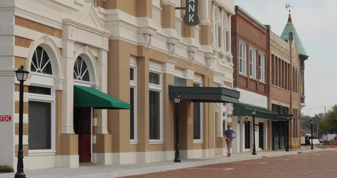 A daytime exterior establishing shot of a generic small town main street business district as a texting male pedestrian walks by. No signage for customization.  	