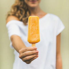 Healthy vegan orange mango citrus ice cream popsicle in hand of young woman with yellow wall at background, square crop. Summer dessert and cheerful summer mood concept