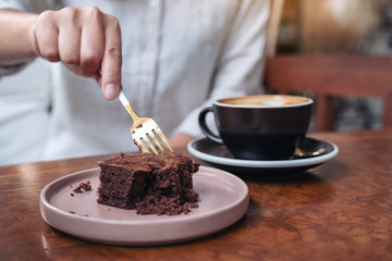 A woman cutting brownie cake with fork with coffee cup on wooden table in cafe