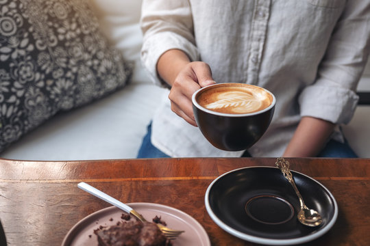 Closeup image of a woman's hand holding and drinking hot latte coffee with brownie cake on the table