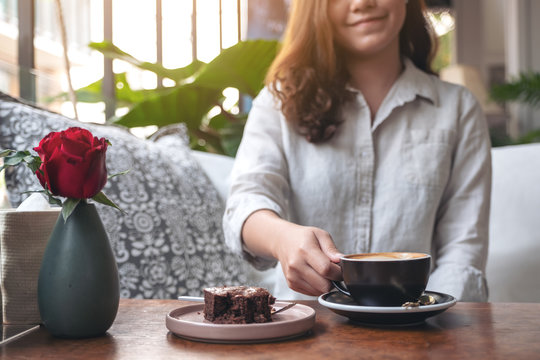 Closeup image of a beautiful woman holding and drinking hot latte coffee with brownie cake on the table in cafe