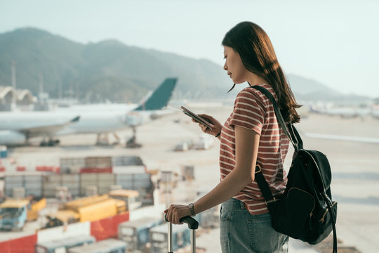 side view of beautiful travel lady with backpack and luggage suitcase walking to departure lounge in hall. tourist woman standing near window using cellphone chatting online airplanes on runway.