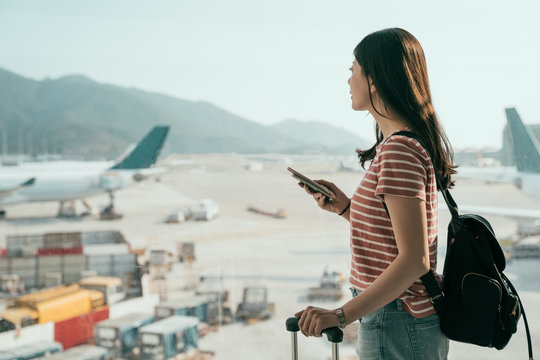 side view of young asian travel woman standing with mobile phone near airport window waiting for flight. beautiful girl with backpack and luggages in hall of lounge area looking out runway airplane