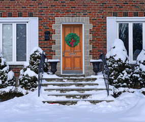 snow covered front steps of brick house with elegant wooden door with Christmas wreath