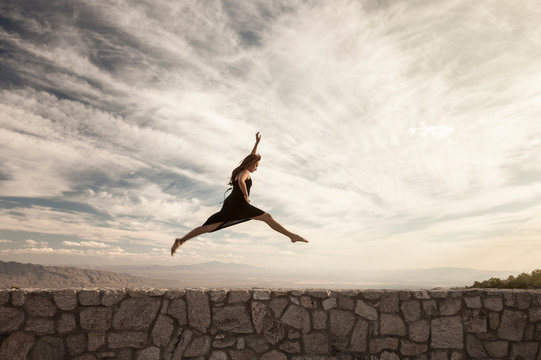 Beautiful, healthy, strong, Hispanic woman leaping jumping over stone wall with dramatic sky background