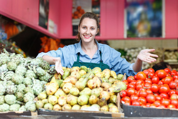 Seller woman is standing near shoecase with vegetables and fruits on the workplace in the market.