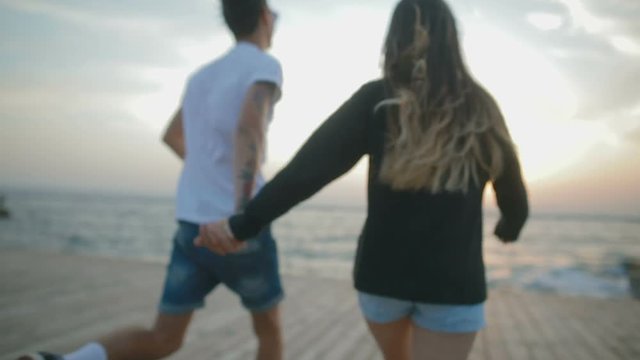 slow motion close backside view brunette girl joining hands with boyfriend runs on boardwalk against sunset over sea