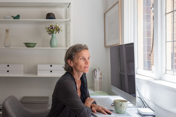 Middle aged woman sitting at home office desk looking happy and hopeful (selective focus)