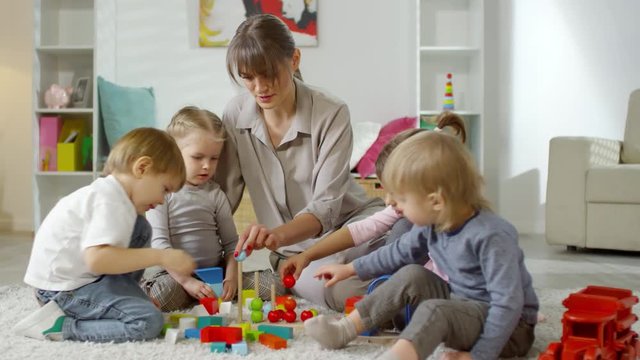 Little children and female teacher sitting on carpet in playroom and playing with colorful educational toy in kindergarten