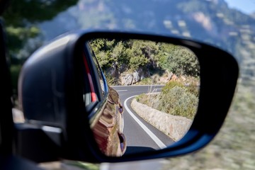  Car driving travel concept - car rear view mirror view of a mountain road with rocks and trees 