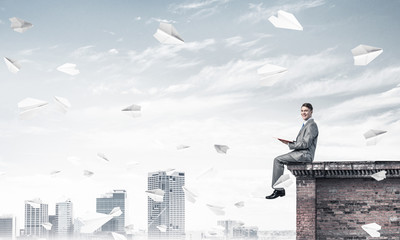 Man on roof edge reading book and cityscape at background