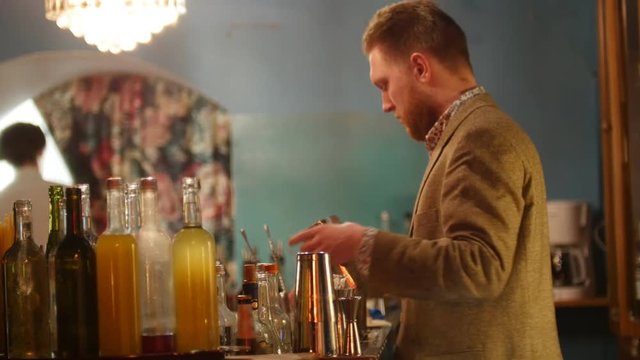 Young professional bartender pouring and shaking a drink and laughing in interior classy bar with soft interior lighting