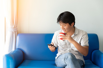 Handsome Asian man using mobile phone while drinking some coffee and sitting on blue sofa at the morning
