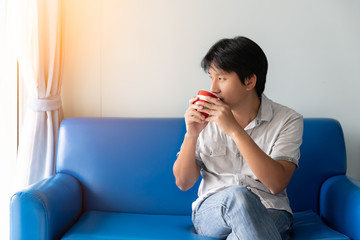 Fototapeta na wymiar A man drinks coffee in the morning while sitting on the blue sofa. He looks at outside and thinks to find creative ideas and inspiration