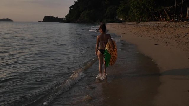 Attractive brunette girl walks on an empty beach with giant inflatable pineapple