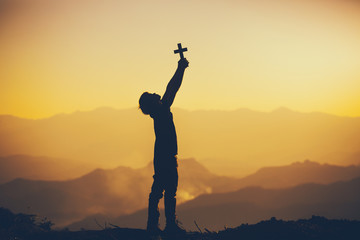 Young man standing holding christian cross for worshipping God at sunset background. christian silhouette concept.