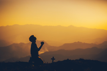 Young man kneeling down and praying with christian cross at sunset background. christian silhouette...