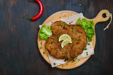 Large Viennese schnitzel on a wooden board with lemon on a dark background. Meat dish. Top view, flat lay.