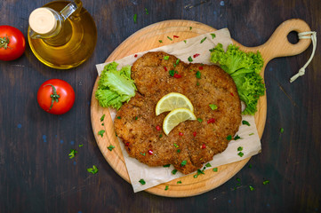 Large Viennese schnitzel on a wooden board with lemon on a dark background. Meat dish. Top view,...
