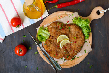 Large Viennese schnitzel on a wooden board with lemon on a dark background. Meat dish. Top view,...